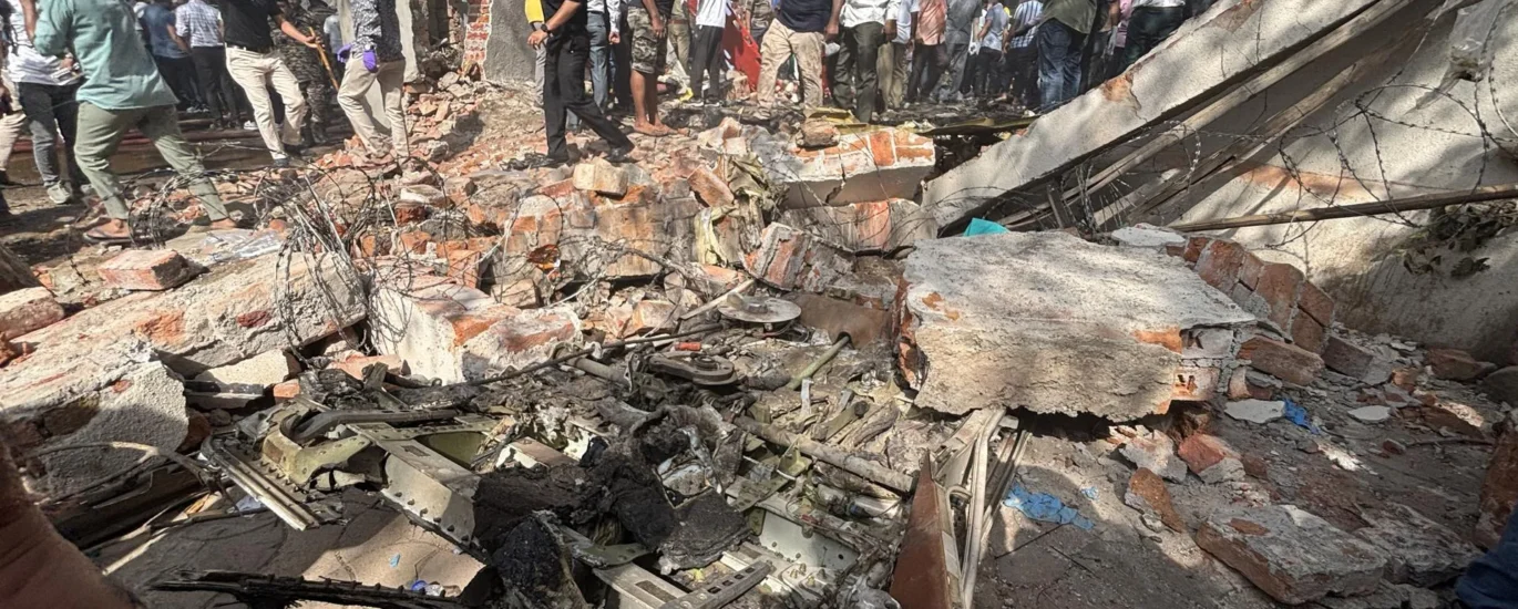 Crowd gathers around rubble and debris of a destroyed building after a plane crash in a residential area, with charred aircraft parts scattered among the wreckage