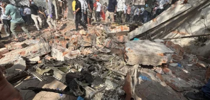 Crowd gathers around rubble and debris of a destroyed building after a plane crash in a residential area, with charred aircraft parts scattered among the wreckage
