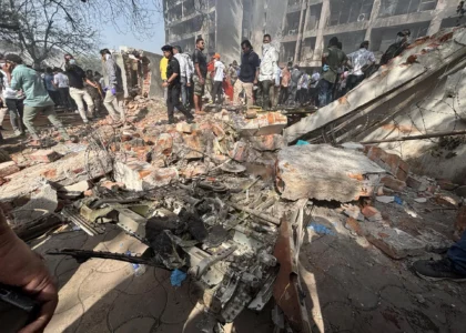 Crowd gathers around rubble and debris of a destroyed building after a plane crash in a residential area, with charred aircraft parts scattered among the wreckage